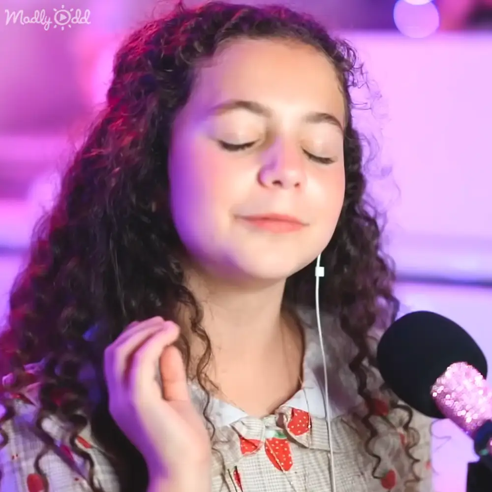 A young adult female wearing a plaid shirt with a bow tie in an indoor performance setting.