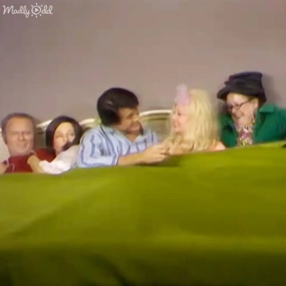 A group of people gathered around a table indoors; adult male with glasses and red shirt, adult female in white garment, young adult male in light blue striped shirt, child with long blonde hair and pink headband, older adult female wearing green top or dress and black hat.