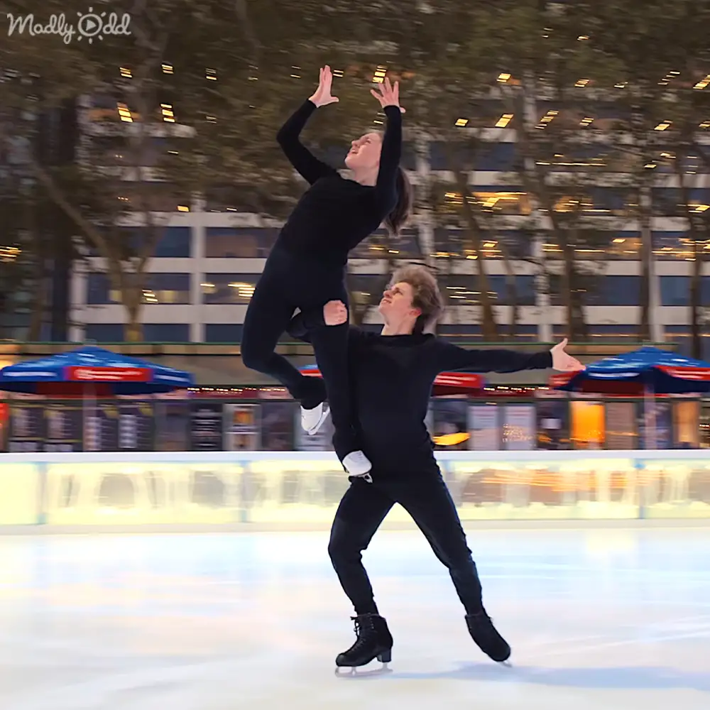 A young adult female figure skater in a black long-sleeve top and pants skating with another young adult male at an indoor ice rink surrounded by seating, umbrellas, and trees.