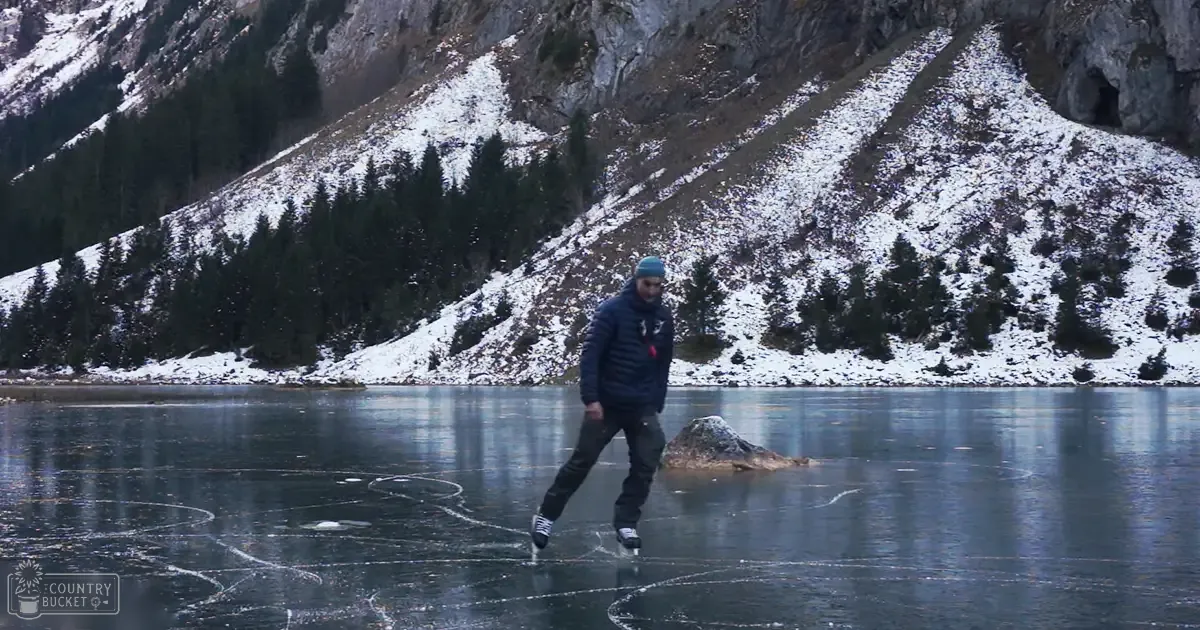 A young adult male skating on a frozen lake in a mountainous, snow-covered region with patches of evergreen trees. The light is natural daylight.