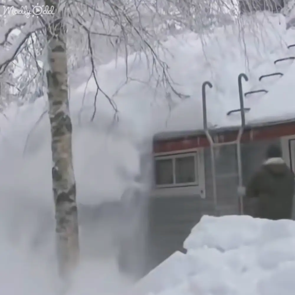 A snow blower clears a residential driveway in winter, with an overcast sky and gray house with red trim.