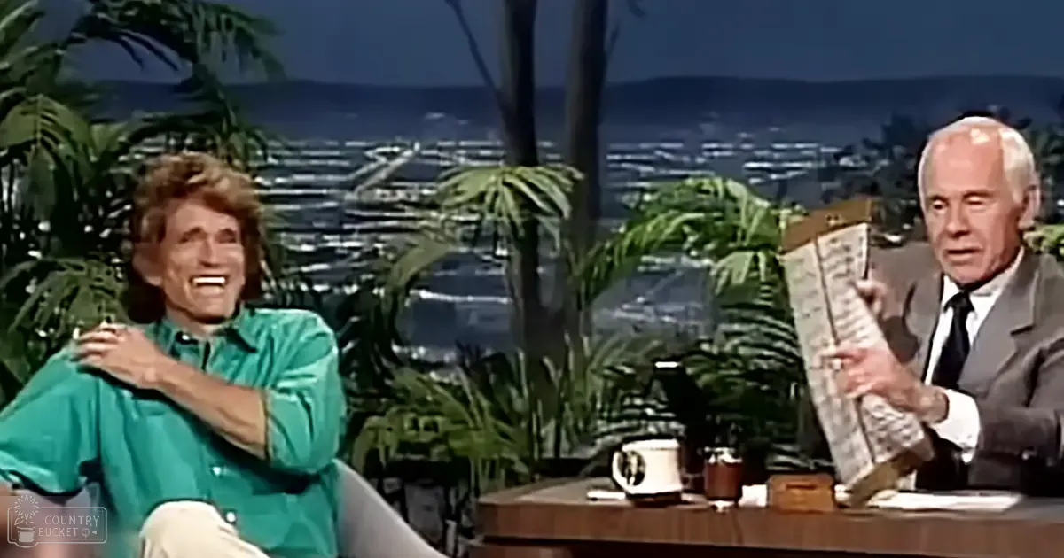 Talk show guest and host on an outdoor studio set with palm trees and a scenic coastal view, natural daylight illuminating the scene.