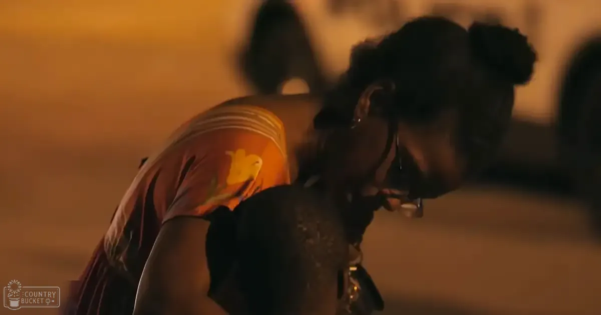 A young adult female wearing a sleeveless yellow floral top and dark pants walks through an outdoor urban area at night under dim orange lighting.