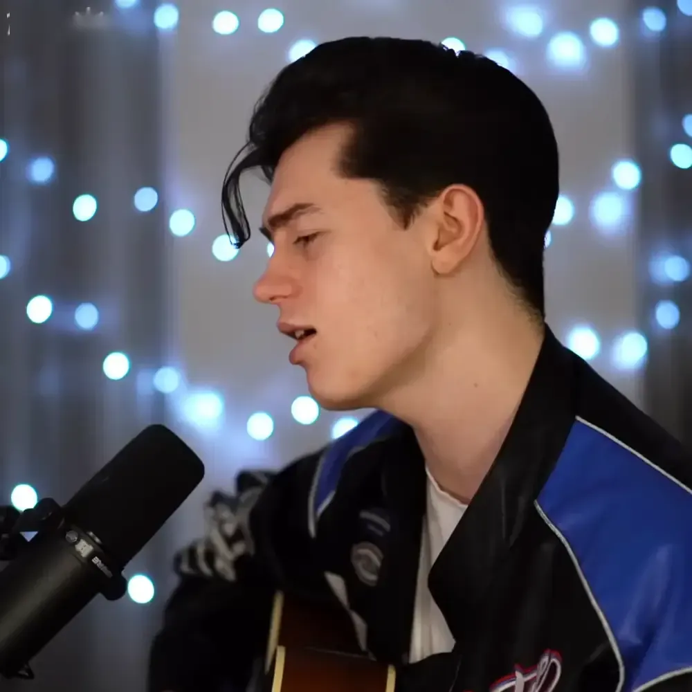 A young adult male singer wearing a black and blue leather jacket with white trim, holding a microphone in an indoor setting illuminated by string lights.