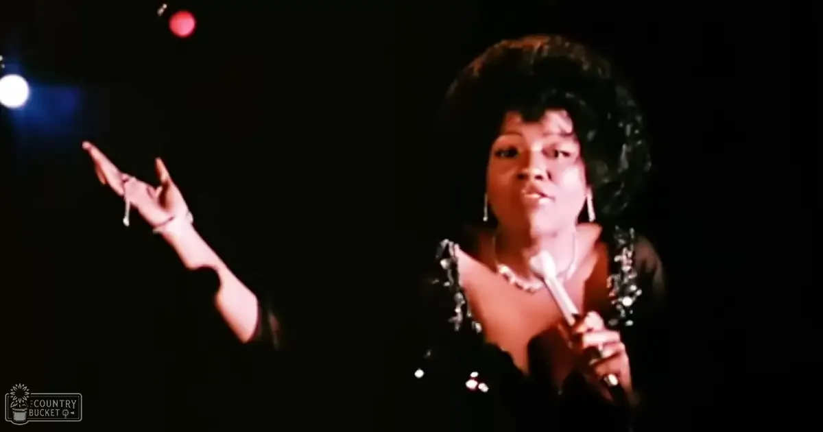 An adult female performer wearing a black, elegant silk or satin garment with a deep neckline and short sleeves, accessorized with dangling earrings and a necklace, holding a microphone on stage under red lighting.
