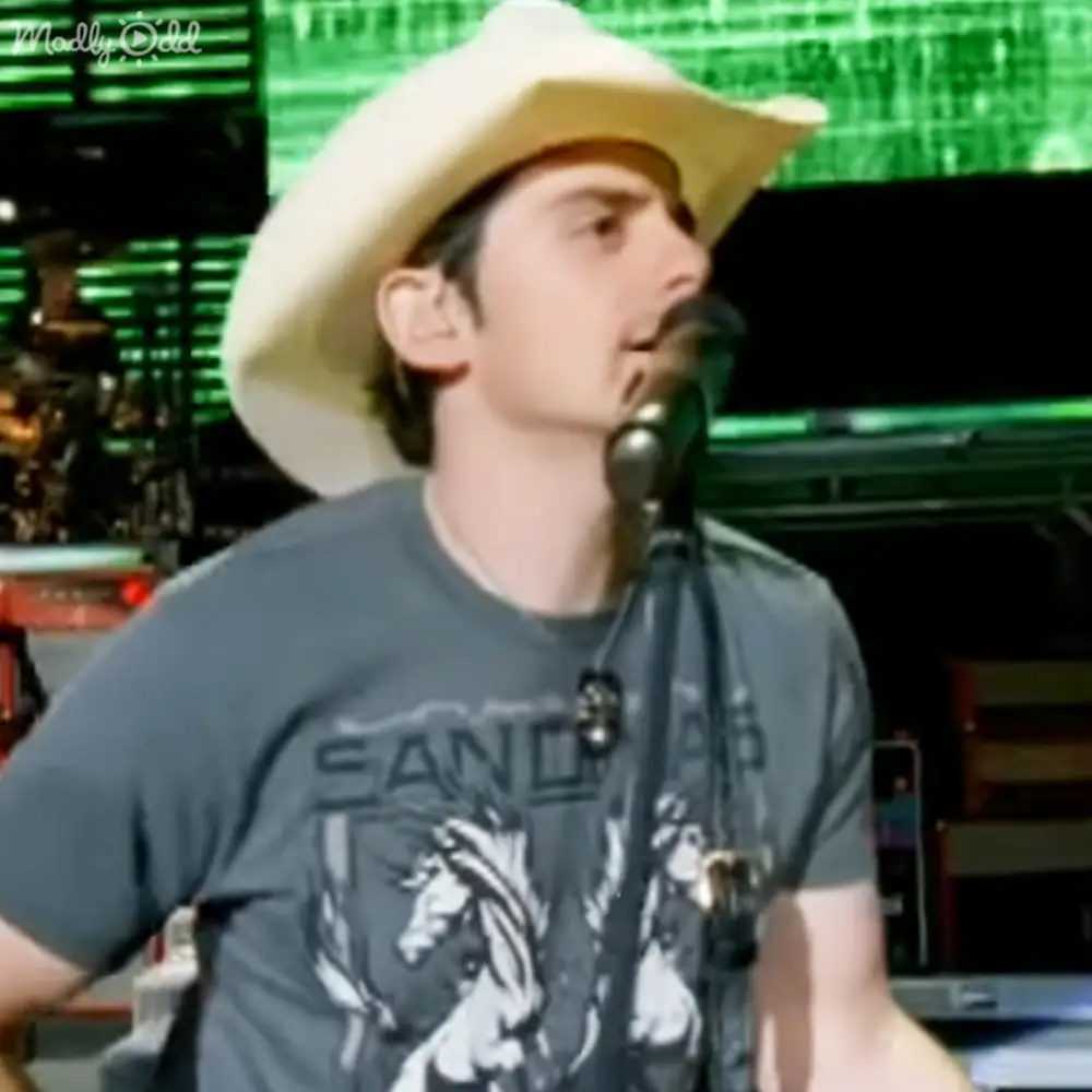 A young adult male wearing a dark gray 'SAND' graphic t-shirt and light-colored cowboy hat stands in an indoor performance setting with stage lighting.