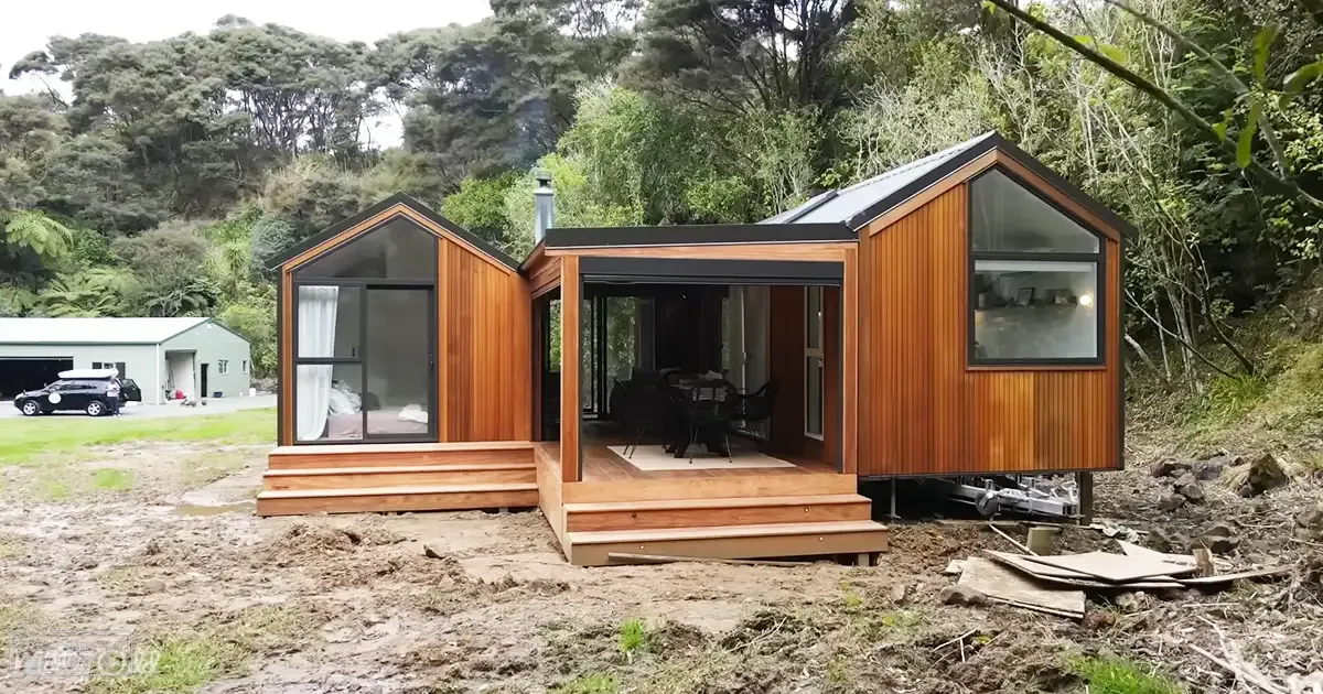 Modern modular home with outdoor seating and vehicles in a rural, forested area during spring or summer.