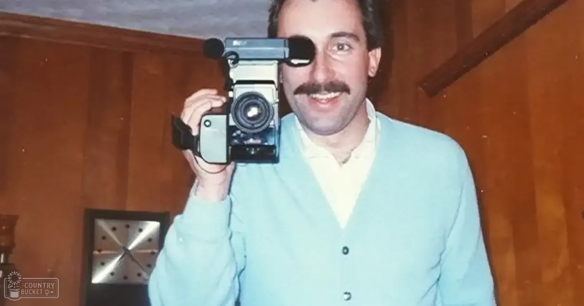 An adult male wearing a light blue cardigan sweater with a collared shirt underneath, holding a vintage camera indoors with wooden paneling and a wall clock visible.