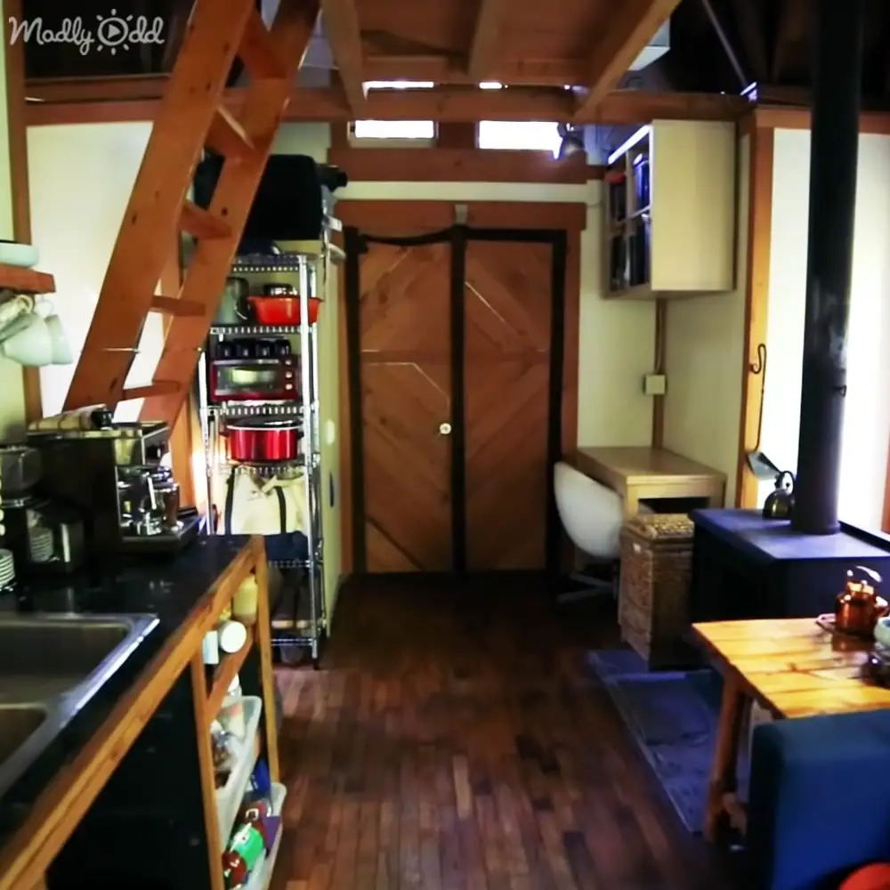 A small, cozy interior space with wooden beams and rustic decor, likely in a cabin or tiny home setting. The natural light suggests it is daytime, possibly morning or afternoon.