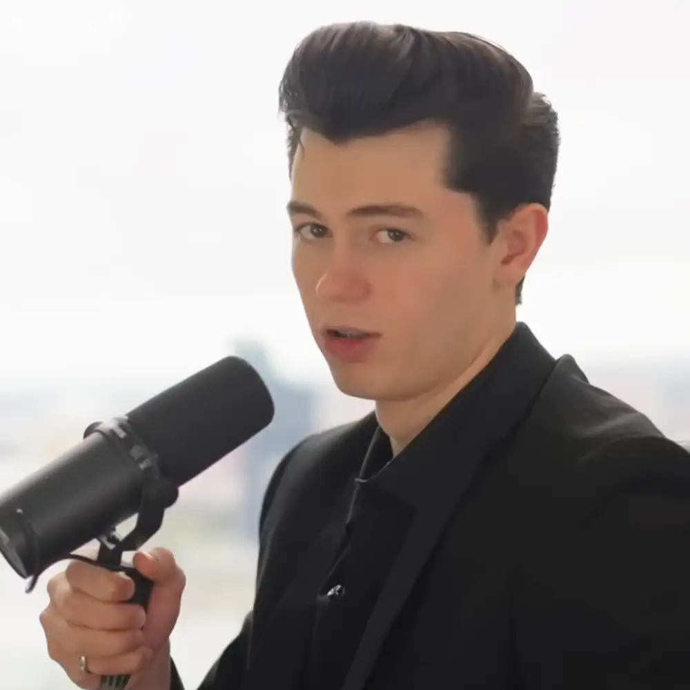 A young adult male wearing a black blazer with a white shirt, holding a professional studio microphone indoors.