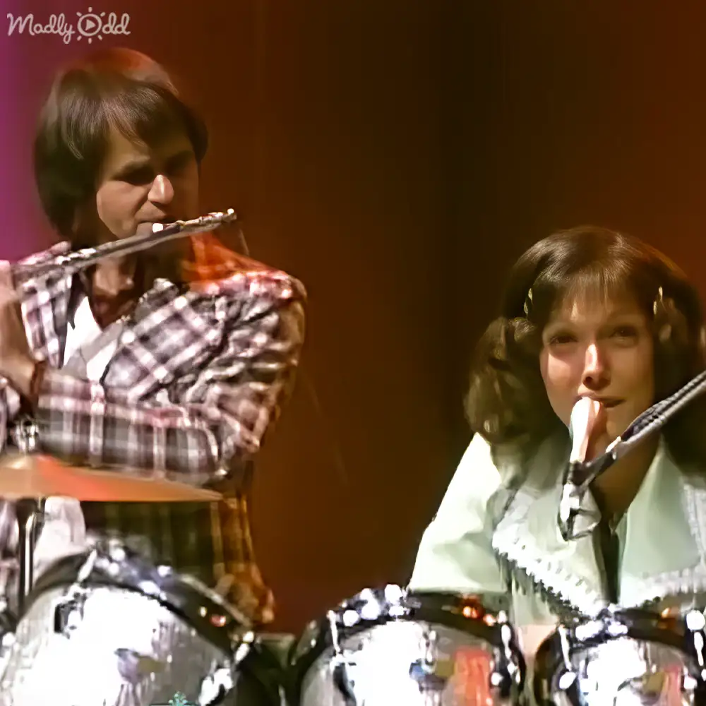Young adult male and female musicians performing with a drum set, wearing plaid shirt and light green jacket respectively, under controlled studio lighting.