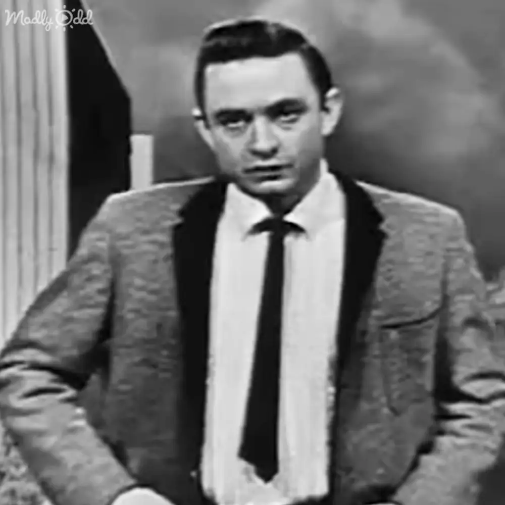 Adult male in a tailored dark suit with white shirt and black tie, standing indoors likely in an office or formal room setting. Background includes striped curtains.