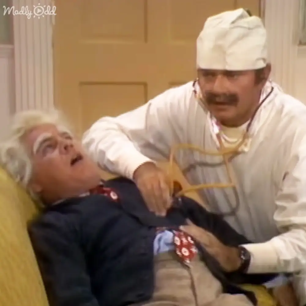 A comedic television show or film scene set indoors with a yellow couch and classic decor elements.