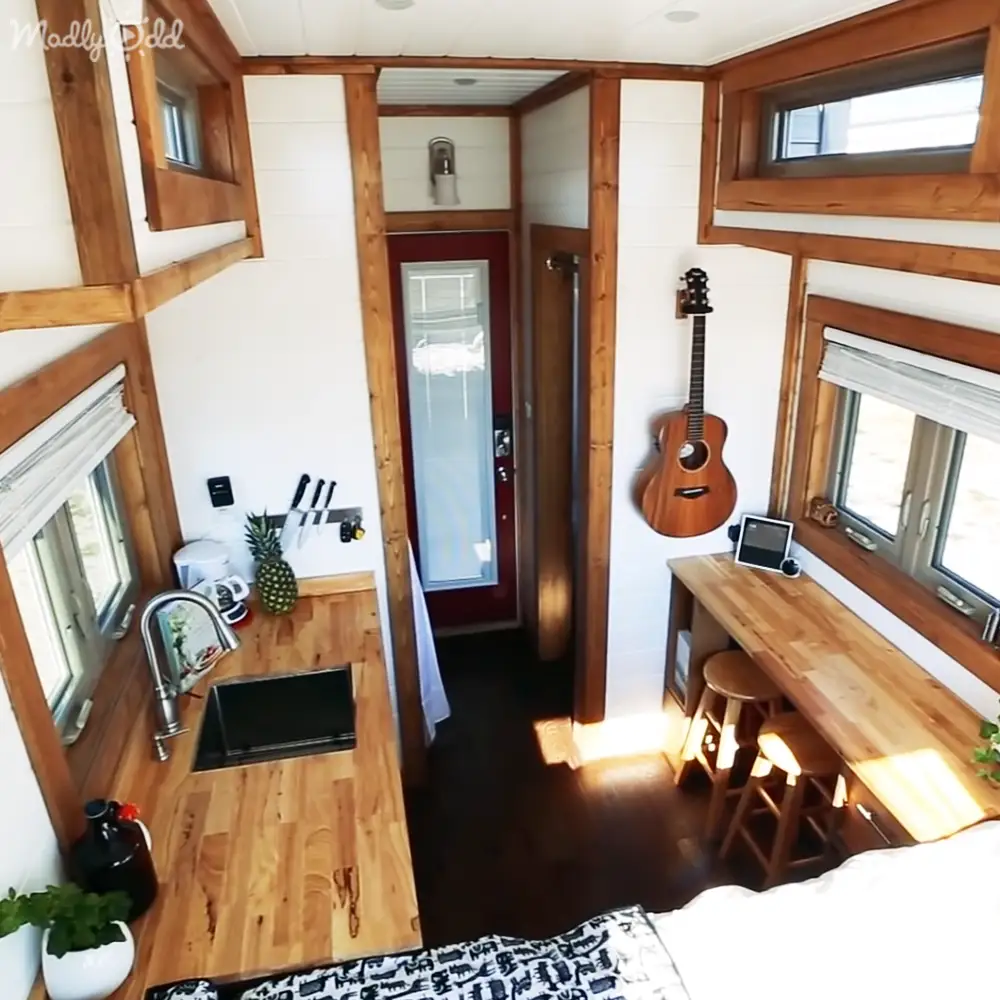 A cozy tiny house interior with a guitar hanging on the wall, a small kitchenette, and various decor items like plants and knives in a magnetic strip.