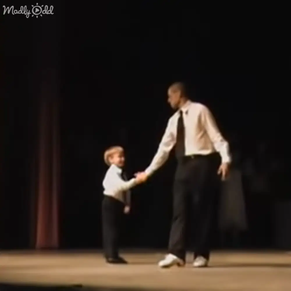 A young boy holding hands with an adult male on a stage in dimly lit performance setting.