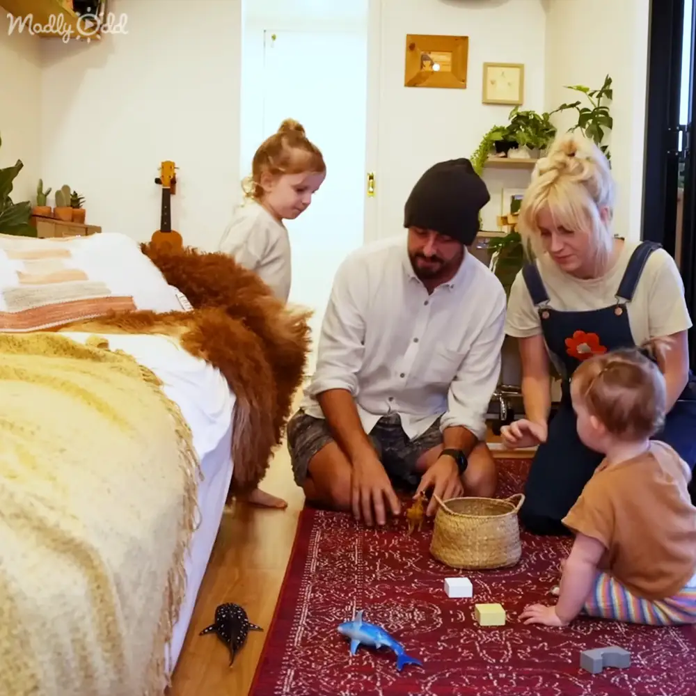 A family of four playing together in a cozy living room with hardwood floors, white walls, and colorful decor. The setting includes toys, a ukulele, and wall art.