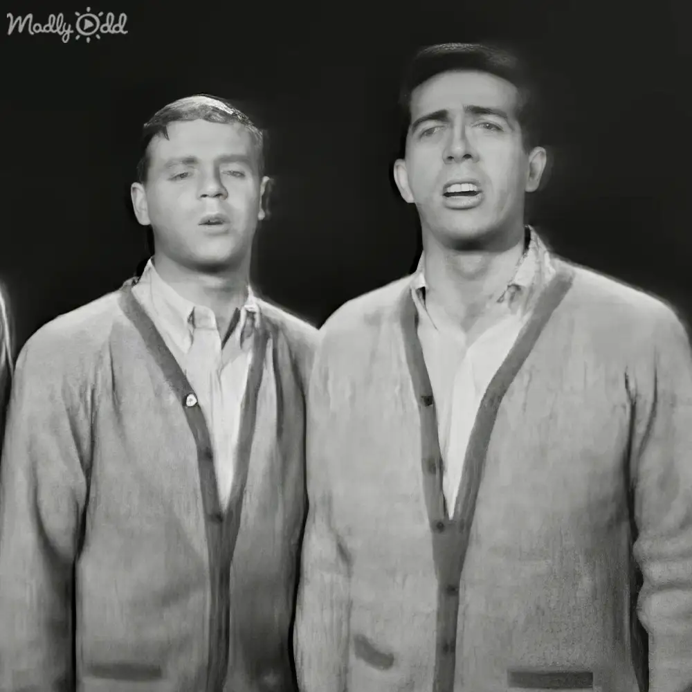 A musical performance scene from the 1960s with two young adult male singers dressed in light-colored button-up shirts and dark cardigans, set against a dark stage backdrop.