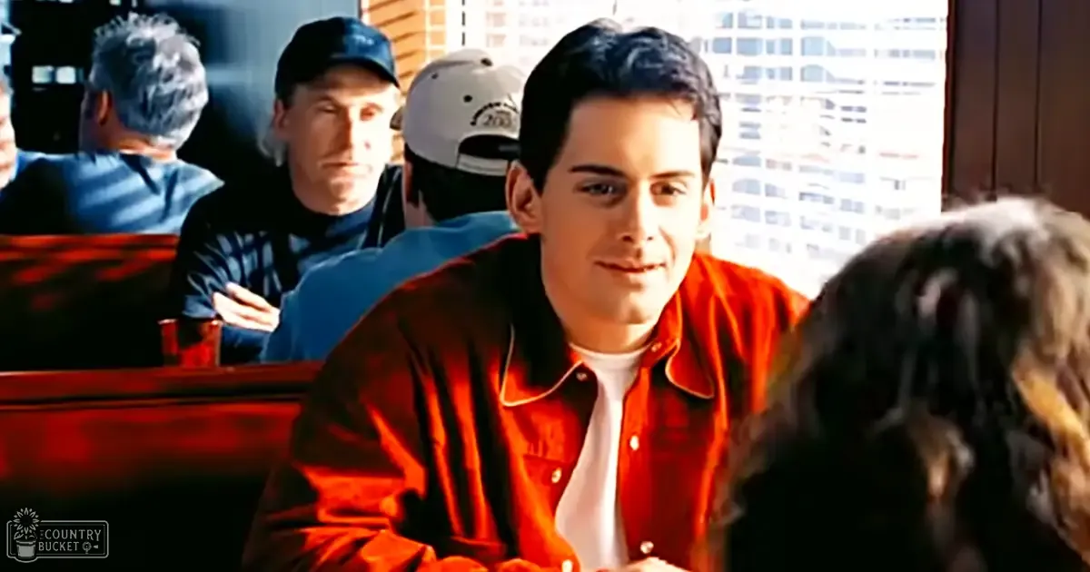 A young adult male in a diner setting, wearing a red jacket and white shirt. Other individuals are present, including middle-aged and older adults, dressed casually.