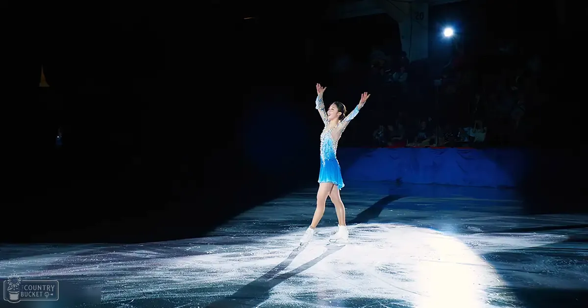 A young adult female figure skater in a blue dress with intricate detailing skating on an indoor ice rink at night under artificial lighting, surrounded by spectators on bleachers.