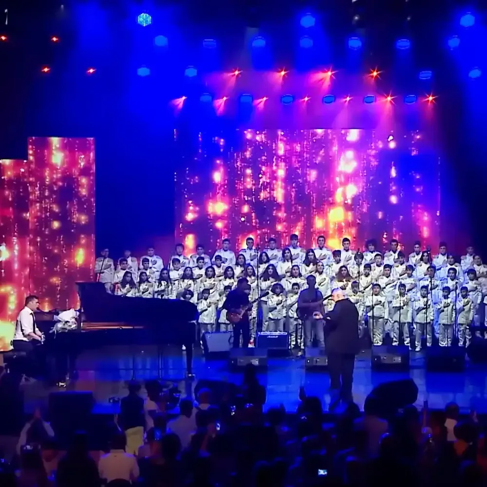 A concert hall scene with a choir and musicians performing in front of an audience, illuminated stage lights creating a festive atmosphere.