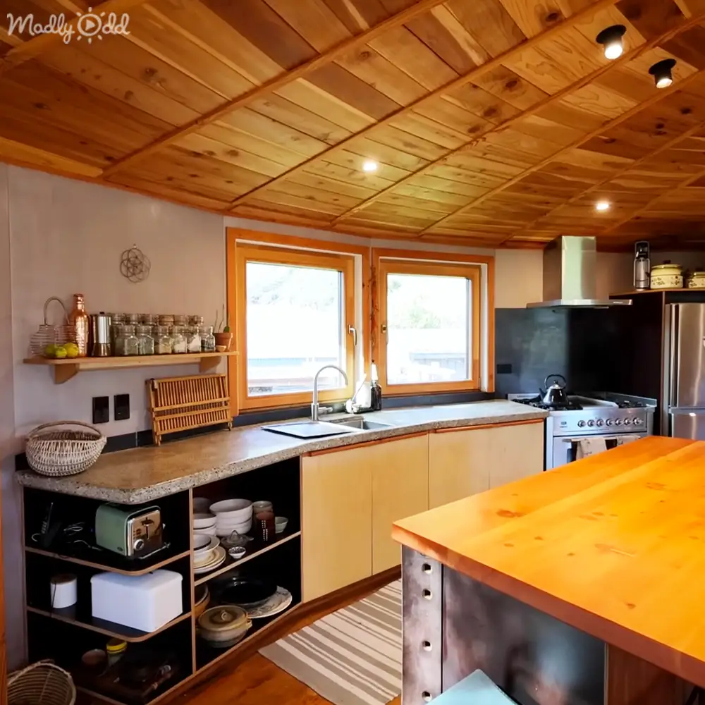 A cozy kitchen scene in a cabin or small home with wooden ceiling, cabinets, granite countertop, stove, refrigerator, open shelving, woven basket, striped rug.