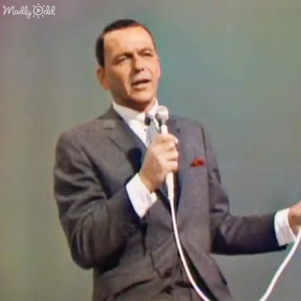 A middle-aged adult male singer wearing a gray suit jacket with a red and black pocket square, performing on an indoor stage in front of an audience.