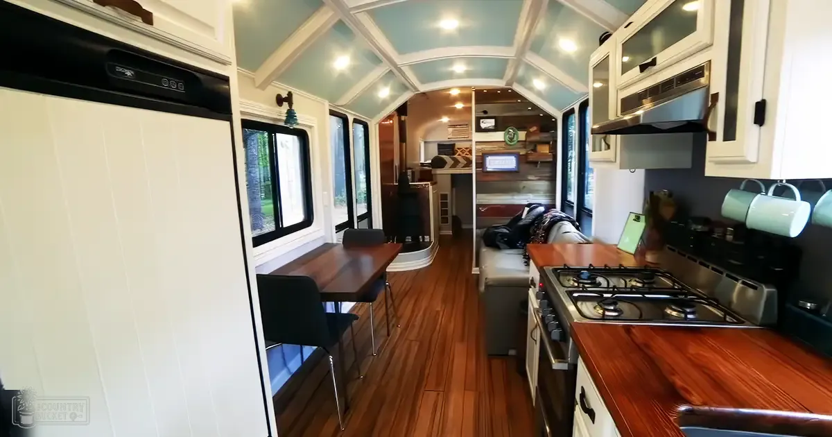 A compact living space with a wooden dining table, white cabinets, gas stove, and natural light from windows.