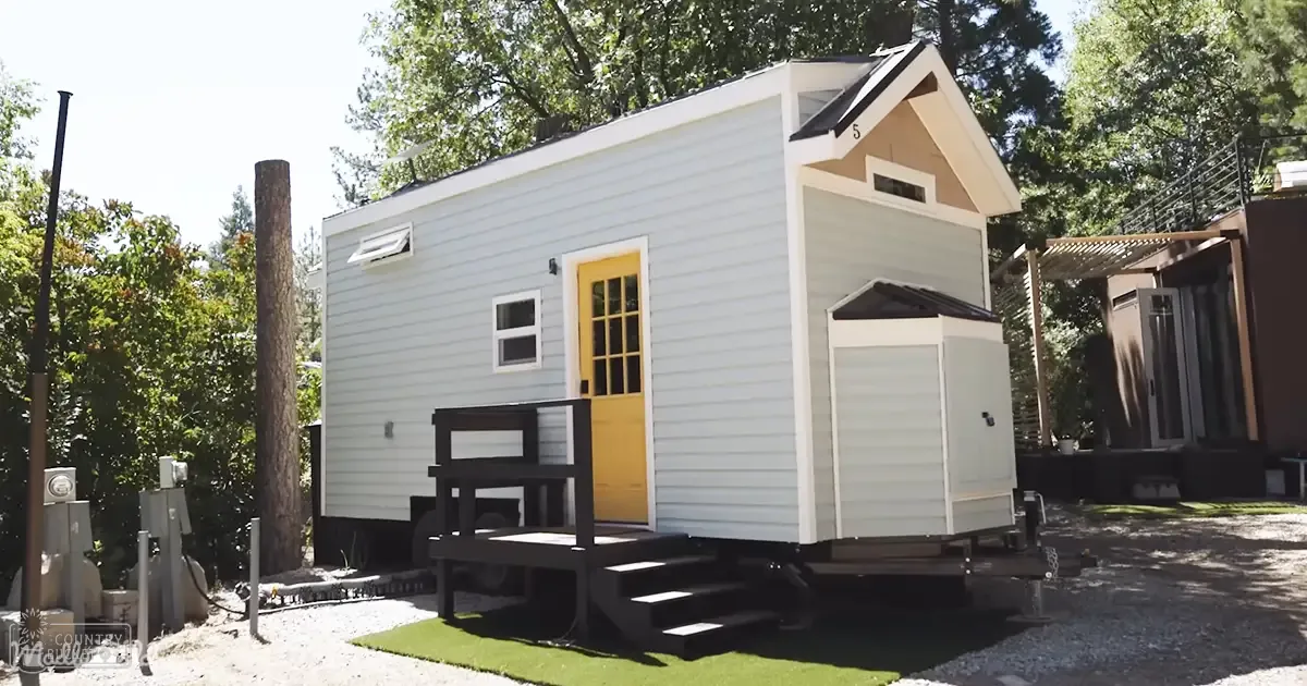 A small white shed attached to a tiny house in an outdoor residential area with gravel ground, trees, and a small wooden table on the porch. The light suggests it's daytime, possibly late morning or early afternoon.