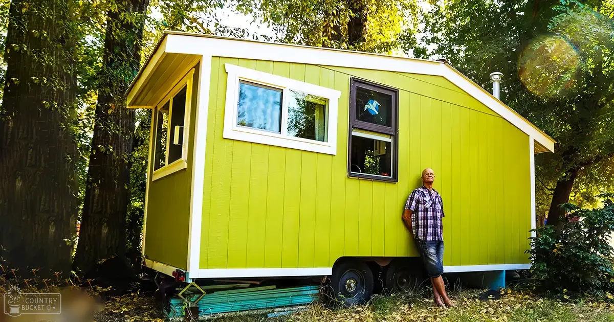 A middle-aged man in a plaid shirt stands next to a small yellow tiny house on wheels, surrounded by tall trees with dappled sunlight filtering through the leaves.