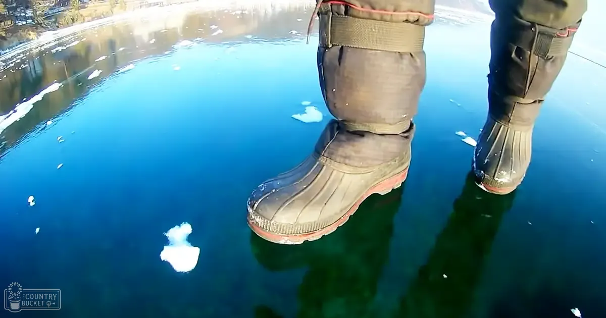 An adult male wearing insulated snow pants and rubber boots stands on ice near a body of water in what appears to be a cold winter setting.