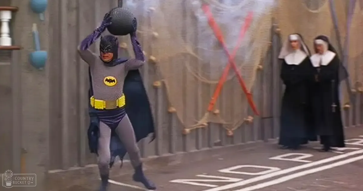 Adult male in a full Batman costume stands indoors, industrial setting with two nuns and ropes hanging from the wall.