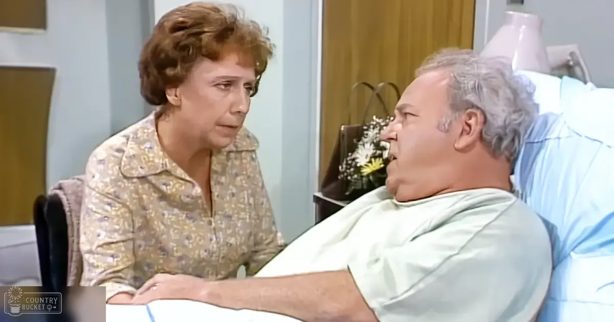 A middle-aged woman in a floral-patterned blouse and an older adult male patient wearing a hospital gown, seated in a room with light blue walls, wooden door, and home decor items like flowers on the bedside table.