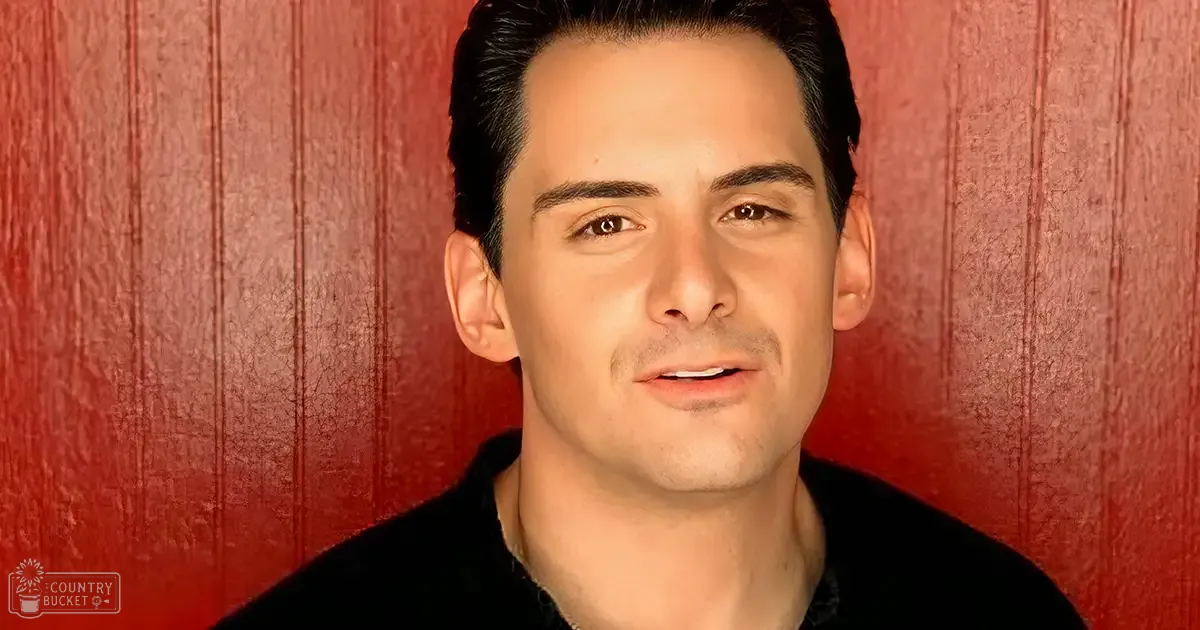 Young adult male in a fitted collared black shirt against vertical red wooden panels, possibly indoors with 'Madly Odd' text visible.