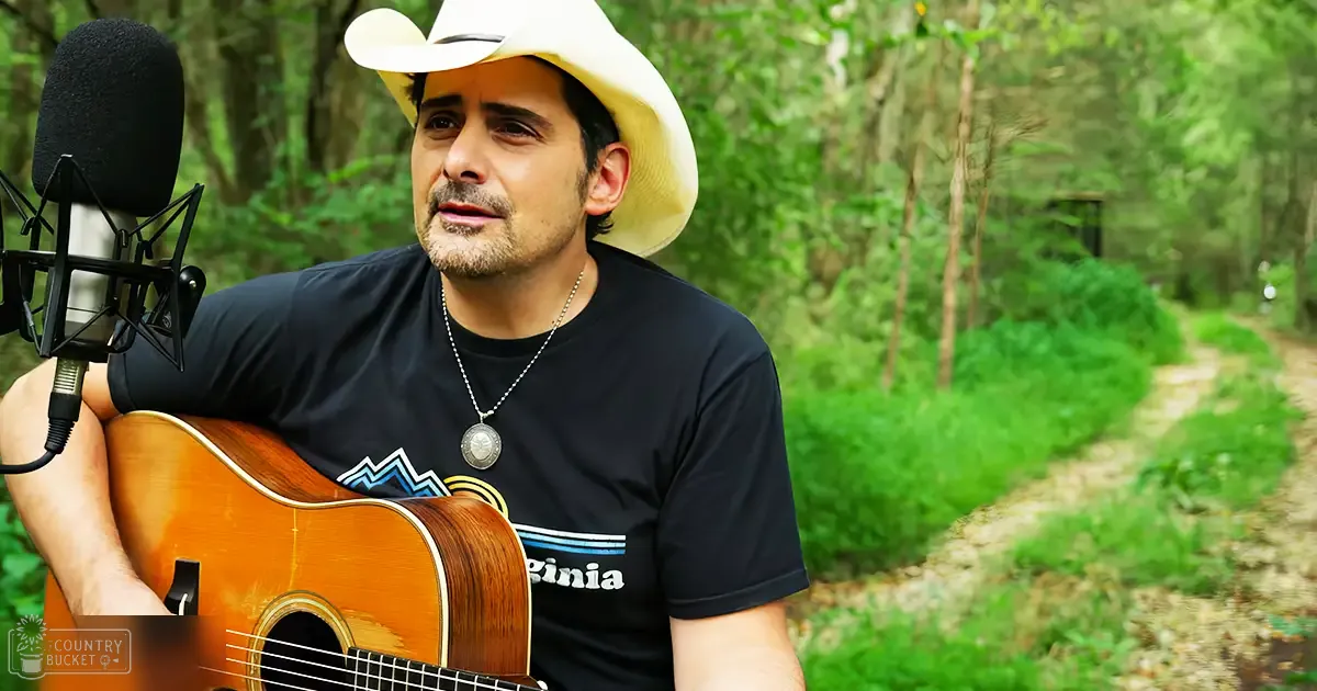 A middle-aged adult male wearing a relaxed-fit, black 'Virginia' t-shirt with mountain graphic design and silver necklace, holding an acoustic guitar outdoors in a forest setting.