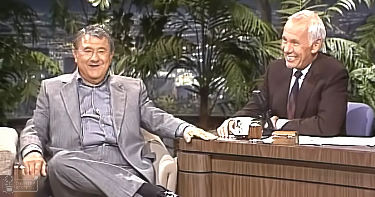 A talk show set with a middle-aged male host seated at a desk, another older adult male seated beside him, both in formal attire; large green plants and an urban backdrop suggest the setting is indoors during daytime.