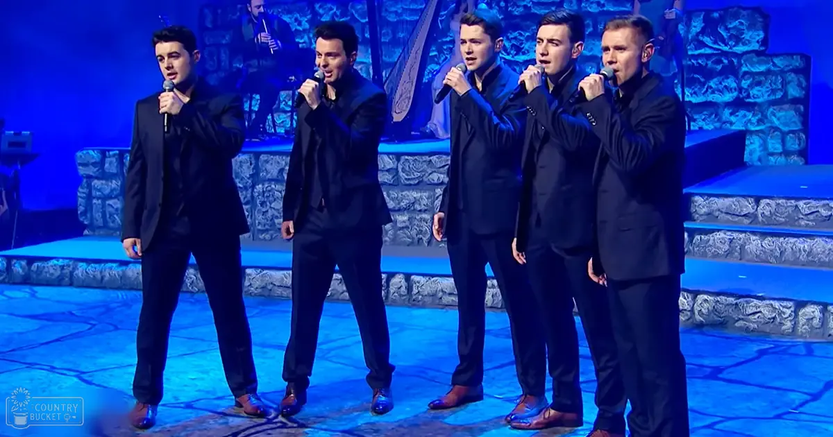 A group of five male singers performing on a stage with an indoor event setting, dressed in black suits under blue artificial lighting.