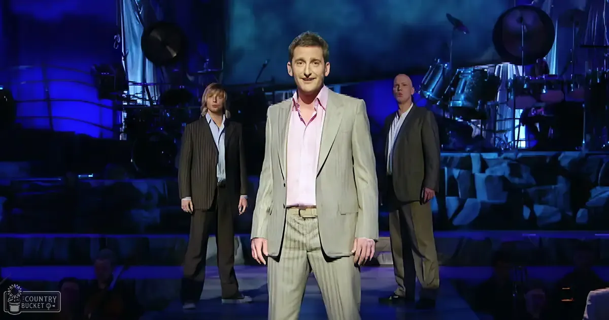 A musical theater performance with an adult male in a light gray suit and another adult male in a dark brown suit on stage, surrounded by musical instruments under dim blue lighting.