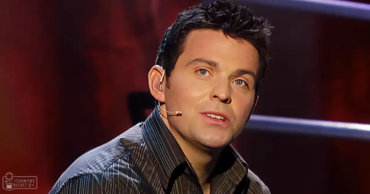 A young adult male wearing a dark brown striped button-down shirt with long sleeves, standing indoors against a backdrop of red curtains.