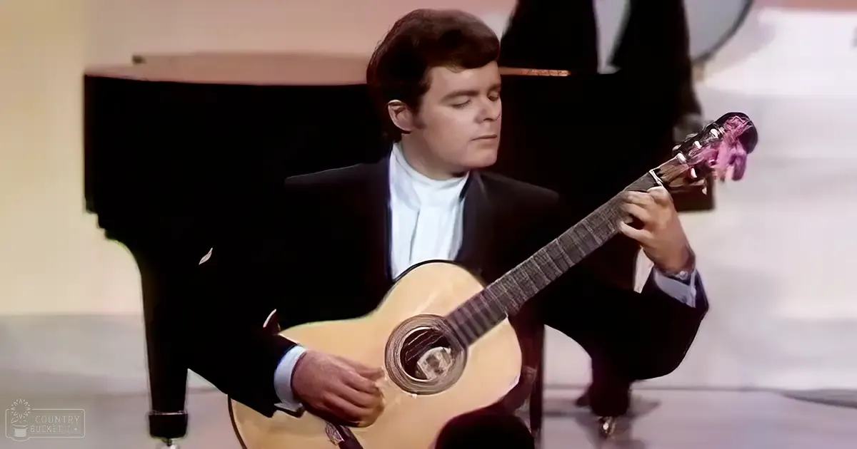 Adult male musician playing an acoustic guitar on stage with formal attire, neutral indoor setting, daytime lighting.