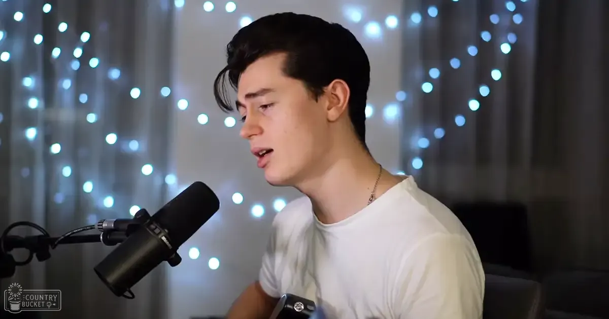 A young adult male wearing a white, relaxed t-shirt with long sleeves stands indoors under string lights, creating a cozy atmosphere.