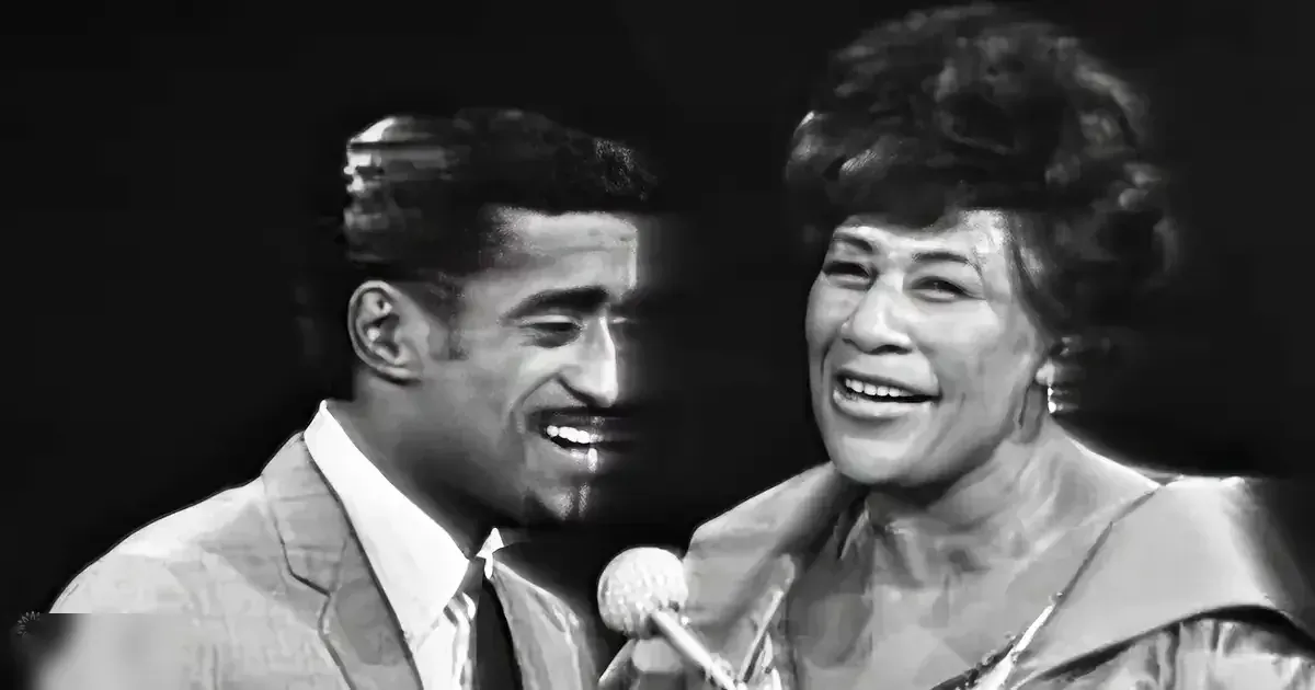 A musical performance scene with two individuals in a dark stage setting, one adult male singer wearing a patterned suit jacket and an older female performer in a light-colored garment with a high neckline.