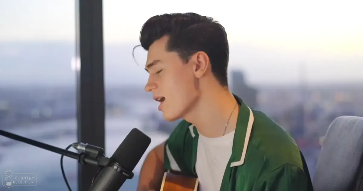 A young adult male singer wearing a green jacket with white trim, singing into a microphone against an urban cityscape backdrop.