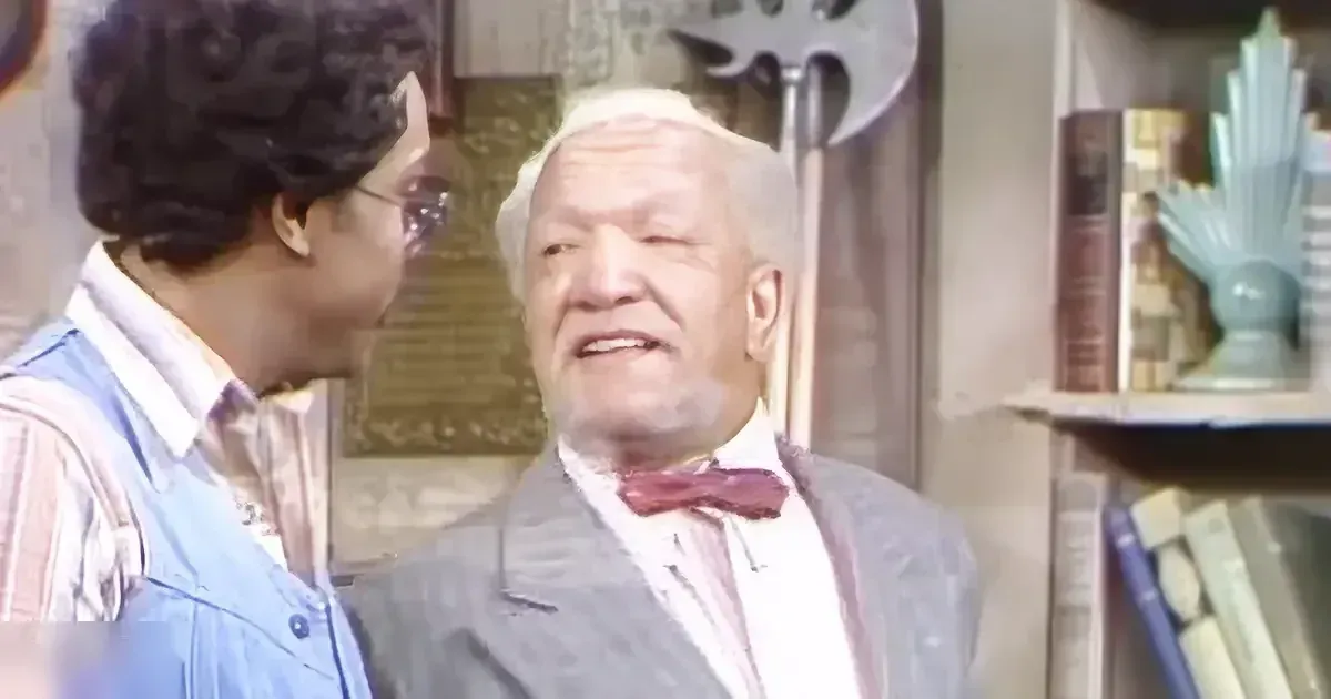 An older adult male in a dark suit jacket with a red bow tie stands beside a child/teen wearing a striped shirt and denim overalls, in a domestic setting with furniture and decor, natural light suggests daytime during spring or summer.