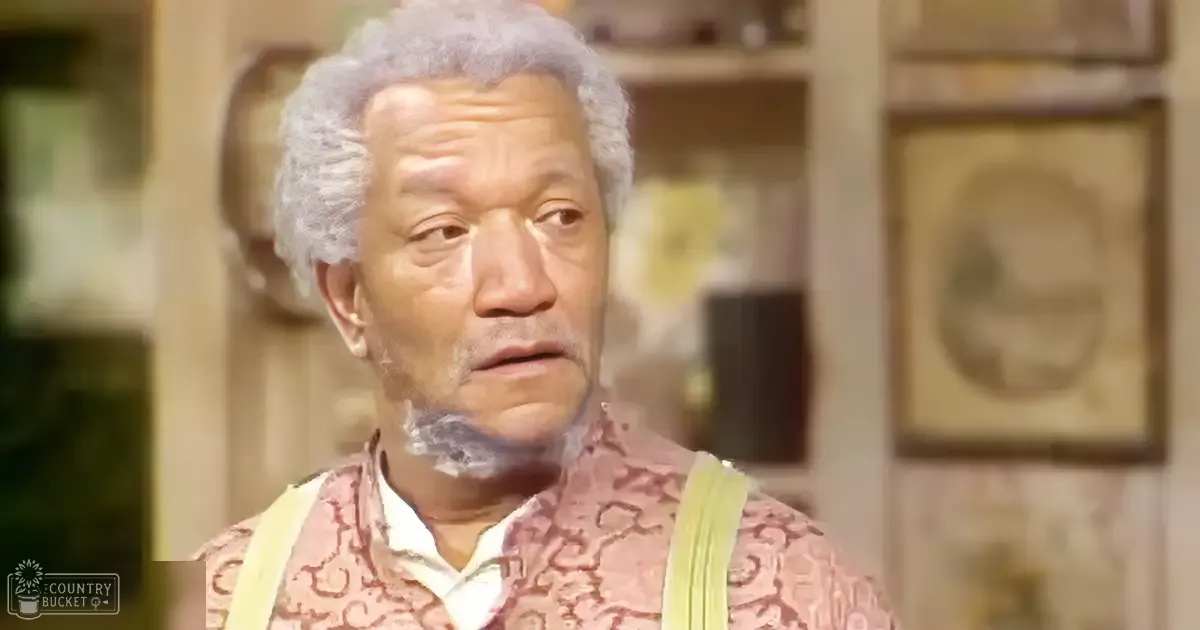 Older adult male wearing a patterned pinkish-red shirt with suspenders, standing indoors in what appears to be a home or living room. The background includes framed artwork and shelves with various items.
