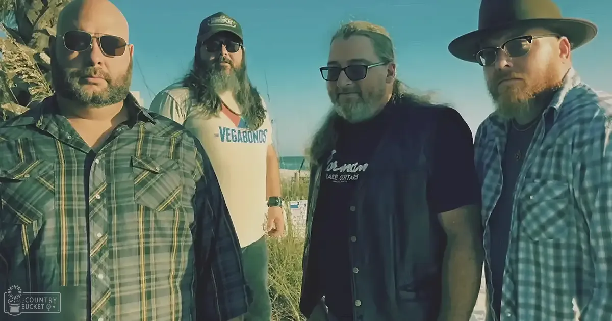 Four men standing outdoors near a sandy area with greenery in the background, wearing casual attire under natural daylight.