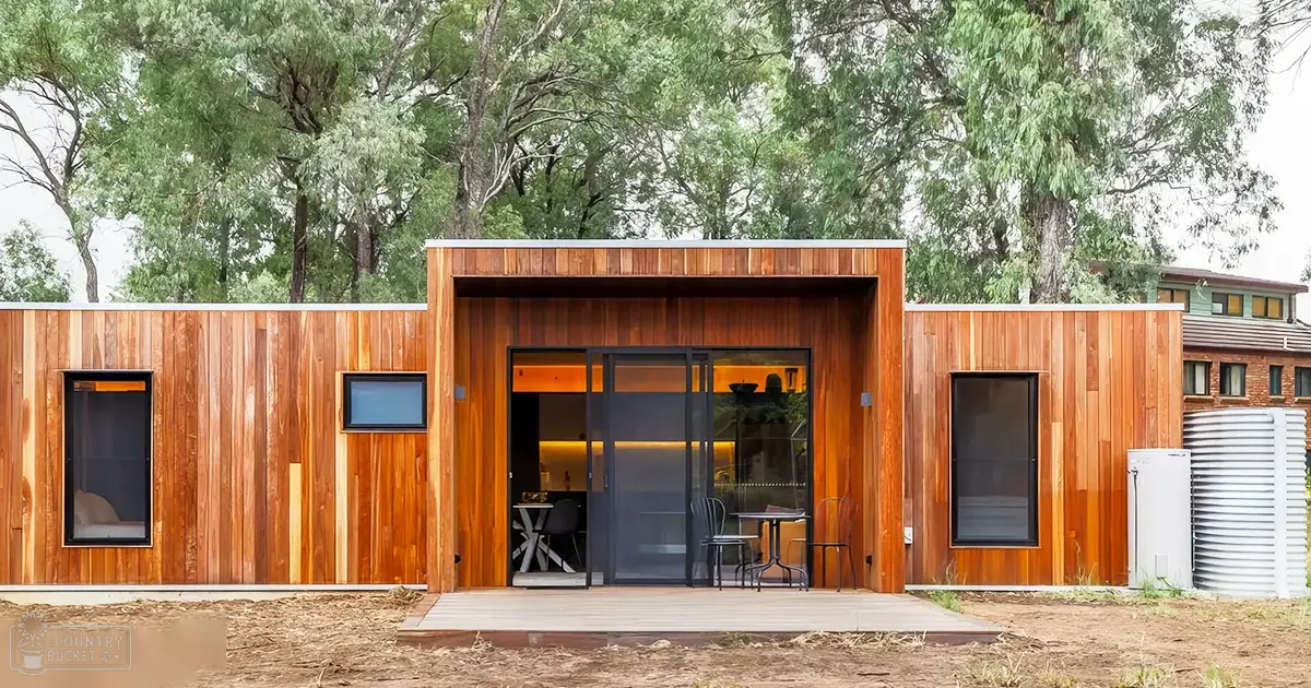 Modern wooden cabin with large glass doors surrounded by lush greenery, suggesting a rural or suburban environment during daytime.