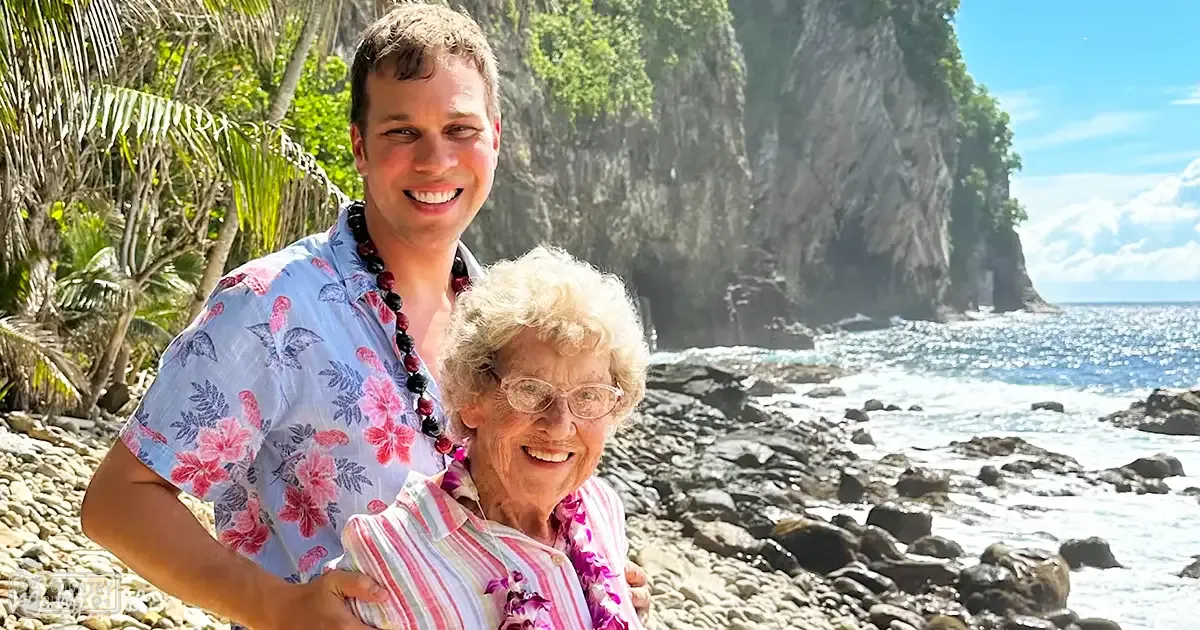 Young adult male in a light blue Hawaiian shirt with pink floral patterns, older adult female in striped shirt by rocky coastline at daytime tropical location.