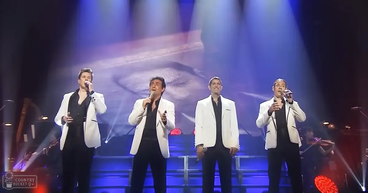 Adult males singing on stage at an indoor concert venue with musical instruments, blue stage lighting, red heart-shaped lights, and a dark backdrop.