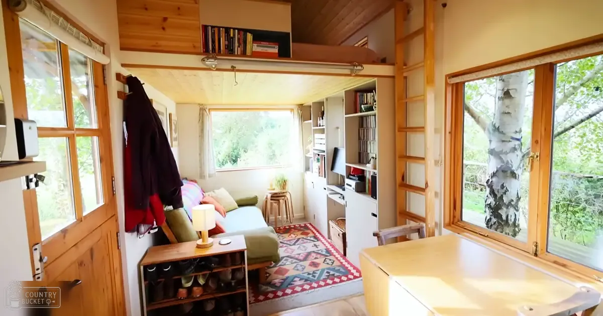 A cozy, compact living space with wooden accents in a cabin or small house, surrounded by lush greenery indicating spring or summer, with earthy tones of brown, beige, and green dominating the scene.