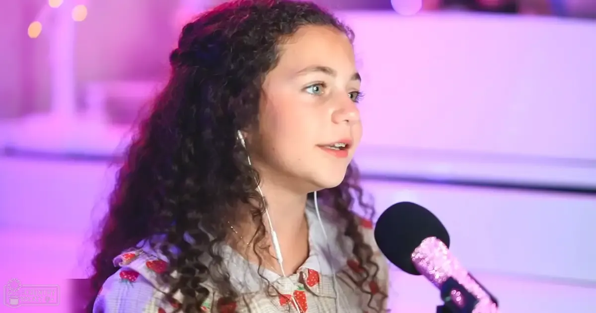A child, likely between 8-12 years old, wearing a patterned light blue top with red strawberry patterns and long sleeves, holding a microphone in an indoor event setting.